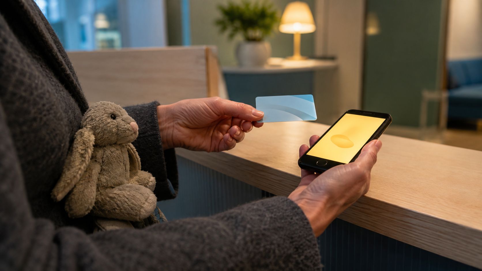 Handen aan de balie van een huisartsenpost in de avond met een zorgpas en een telefoon, en een knuffel onder de arm